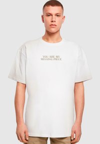Hvid bomulds t-shirt med korte ærmer, med teksten i lys beige, præget "DU ER MIT MANGLENDE PUZZLESTYKKE" i midten.