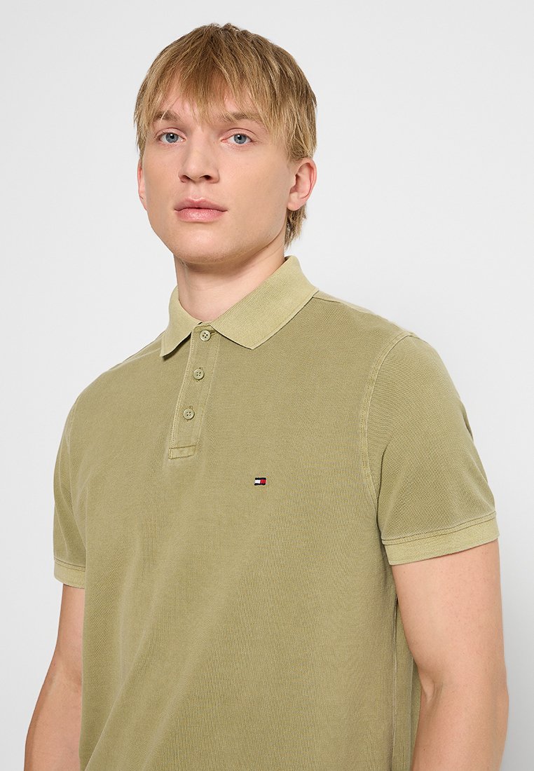 Polo verde oliva hecho de tela texturizada, con una tapeta de tres botones y un pequeño logo en el lado izquierdo del pecho. Mangas cortas.