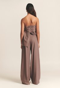 Rjav kombinezon brez ramen z nabrano podrobnostjo na trupcu in širokimi hlačnicami, narejen iz gladkega blaga, z zadrgo na hrbtu in stranskimi žepi.