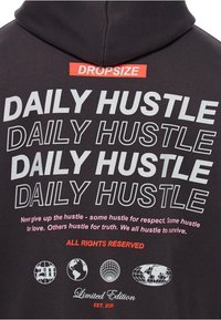 Grauer Hoodie mit auffälligem weißen Text "DAILY HUSTLE" in verschiedenen Größen, roten Akzenten und Globusgrafiken. Weiches Baumwollmaterial, normale Passform.