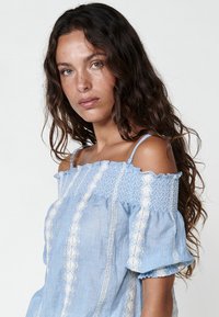 Top azzurro chiaro scoperto sulle spalle con bordo elastico, caratterizzato da motivi ricamati bianchi e maniche a sbuffo. Realizzato in tessuto leggero.