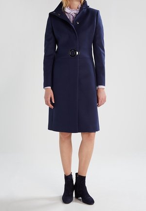 Femme portant un manteau bleu marine mi-long avec un col montant, une ceinture à la taille, un chemisier rose clair et des bottines noires, debout sur un fond blanc.