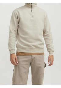 Felpa beige chiaro con zip realizzata in tessuto morbido, dotata di colletto alto e polsini a coste, indossata sopra una camicia marrone.