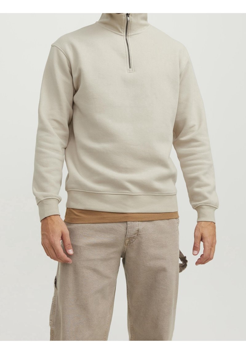 Felpa beige chiaro con zip realizzata in tessuto morbido, dotata di colletto alto e polsini a coste, indossata sopra una camicia marrone.