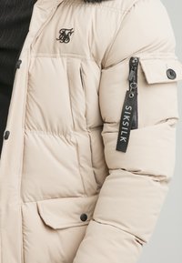 Beige Daunenjacke mit einer Reißverschlusstasche am Ärmel, einem Logo auf der Brust und einem Kunstfellbesatz an der Kapuze. Strukturierter Stoff.