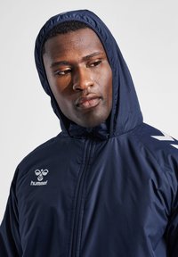 Chaqueta con capucha de color navy con cierre de cremallera, presenta un logo blanco y un diseño en forma de chevron en las mangas. Textura suave, tejido ligero para mayor comodidad.