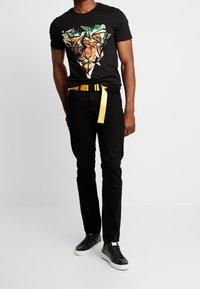 Camiseta negra de manga corta con un gráfico de león geométrico; combinada con jeans negros y un cinturón amarillo. Completa con zapatillas negras.