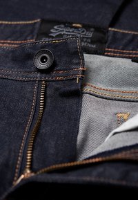 Tummansiniset farkut oransseilla saumoilla, joissa metallivetoketju, kangasmerkki, jossa lukee "Superdry", ja sileä sisävuori.