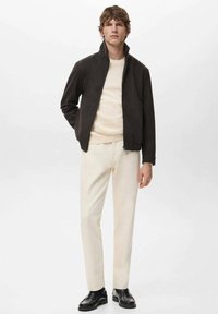 Tmavohnedá bunda na zips z mäkkej látky, skombinovaná so svetlo béžovým svetrom a krémovými nohavicami. Čierne topánky dopĺňajú vzhľad.