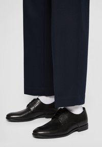 Scarpe eleganti in pelle nera con finitura liscia e design con stringhe, indossate con calzini bianchi, sotto pantaloni blu scuro.