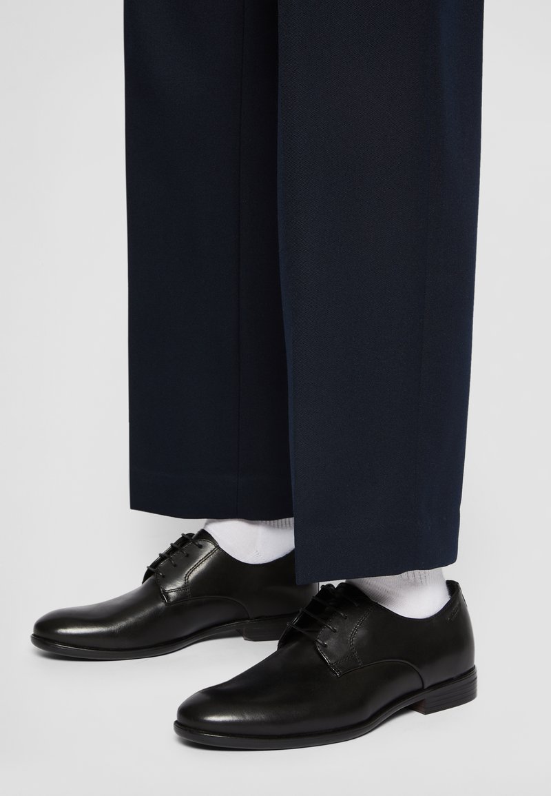 Scarpe eleganti in pelle nera con finitura liscia e design con stringhe, indossate con calzini bianchi, sotto pantaloni blu scuro.
