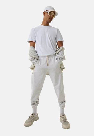 Sportoutfit bestehend aus einem weißen T-Shirt, grauen Sweatpants mit Kordelzug und beigen Turnschuhen. Die Accessoires umfassen eine weiße Baseballmütze und übergroße Brillen.