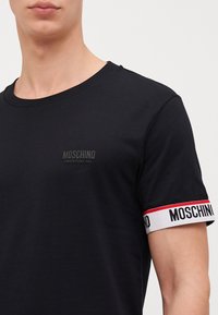 Melna kokvilnas T-kreklu ar apaļu kaklu, piedurknēs ir kontrastējoša lentes ar tekstu, un uz krūtīm ir mazs izcelts "Moschino Underwear" logotips.