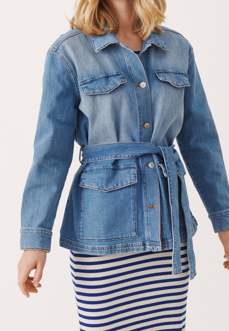 Femme portant une veste en jean bleue ceinturée par-dessus une jupe blanche à rayures bleues, avec des cheveux blonds lâchés visibles au-dessus du col.