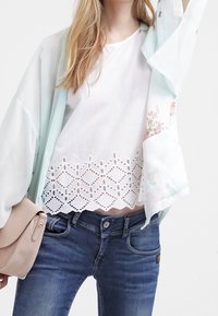 Leichter mintgrüner Kimono mit floralen Akzenten, kombiniert mit einem weißen Oberteil mit Spitzenabschluss und blauen Jeans aus Denim. Inklusive einer beigen Handtasche.