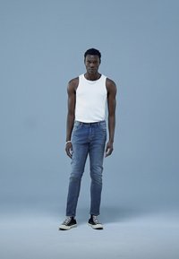 Top fără mâneci alb, blugi din denim albastru, adidași negri cu glezna înaltă. Fundal neutru, model în picioare cu brațele pe lângă corp.