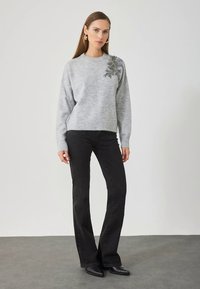 Grijze trui met een gestructureerde afwerking, voorzien van een bloemenapplicatie op de linker schouder. Gecombineerd met zwarte uitlopende broek en laarzen.