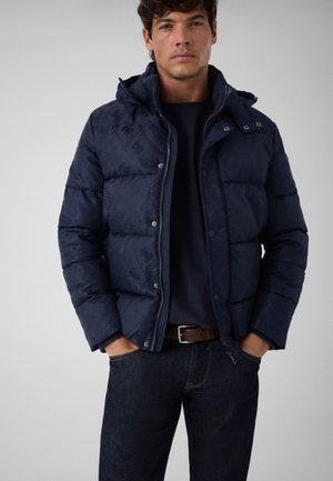 Homme portant une veste matelassée bleu marine avec capuche, un jean foncé et une ceinture marron, debout les mains dans les poches devant un fond uni.