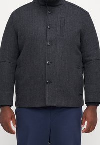 Dunkelgraue Fischgrätjacke mit hohem Kragen, fünf Knöpfen, zwei Fronttaschen und einer dezenten Textur. Mit einer aufgesetzten Brusttasche.