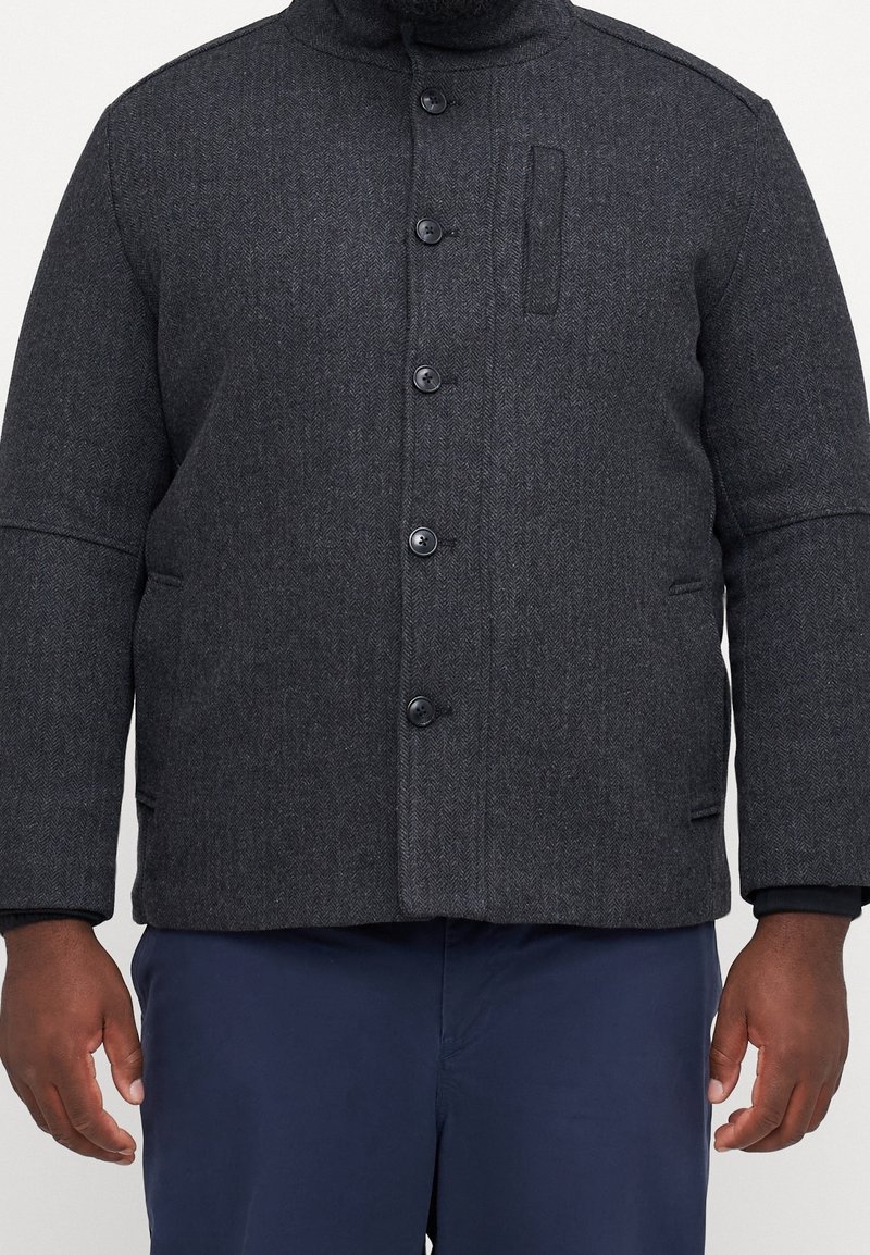 Dunkelgraue Fischgrätjacke mit hohem Kragen, fünf Knöpfen, zwei Fronttaschen und einer dezenten Textur. Mit einer aufgesetzten Brusttasche.