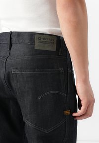 Dark denim jeans with contrasting yellow stitching, G-Star label on back, and angled pockets. Features a fitted design and durable material.