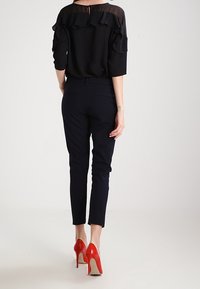 Černá blůza s vrstvenými volány a průsvitným vrškem, kombinovaná s úzkými námořnickými kalhotami a červenými lodičkami na vysokém podpatku.