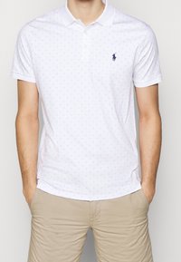 Witte polo shirt met lichtblauw stippenpatroon, korte mouwen en een drieknoopsluiting. Beschikt over een klein logo op de linkerborst.