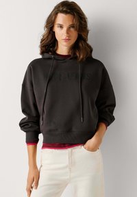 Felpa con cappuccio grigio scuro con logo "Pepe Jeans", polsini a costine e vestibilità comoda. Indossata sopra una maglia color bordeaux e abbinata a jeans chiari.