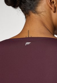 Burgundy shirt with a smooth texture and a wide neckline, featuring a small white logo at the back of the neck.