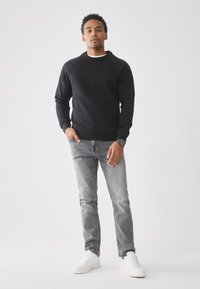 Man staat met één hand in zijn zak, gekleed in een zwart sweatshirt, grijze jeans, witte sneakers en een polshorloge tegen een effen lichte achtergrond.