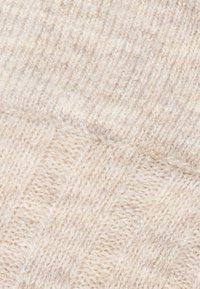 Světle béžová pletená textilie s žebrovaným a diagonálním vzorem. Měkká textura s variací tloušťky příze, vytvářející hloubku a zajímavost.