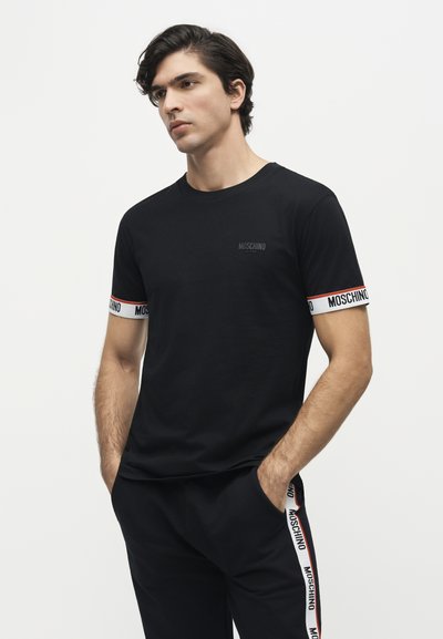 Homme portant un t-shirt noir Moschino et un pantalon avec des bandes à logo sur les manches et les côtés du pantalon, debout les mains dans les poches devant un fond blanc.
