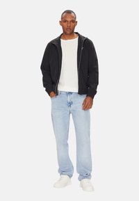 Giacca nera con zip sopra una t-shirt bianca, abbinata a jeans azzurri e sneakers bianche. Design semplice, vestibilità casual e senza motivi visibili.