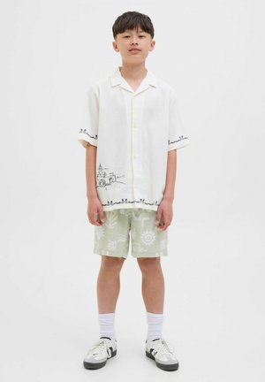 Kortärmad, vit skjorta med knappstängning och grafisk design vid nederkanten; matchas med ljusgröna shorts med vita abstrakta mönster.