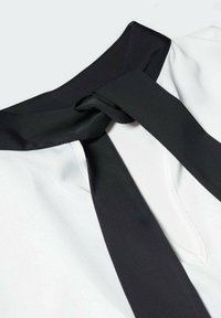 Blouse blanche avec un col en sangle noir, dotée d'un détail de nœud. Le tissu semble lisse avec une texture soyeuse, mettant en valeur le contraste des couleurs.