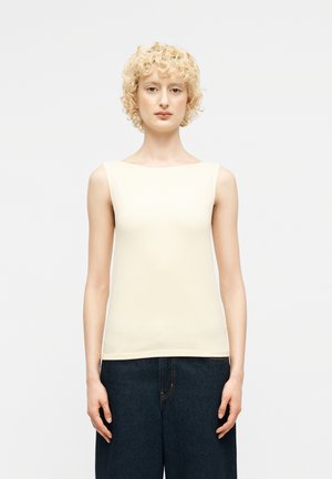 Personne aux cheveux blonds courts et bouclés portant un haut sans manches couleur blanc cassé et un jean large foncé, posant devant un fond clair uni.