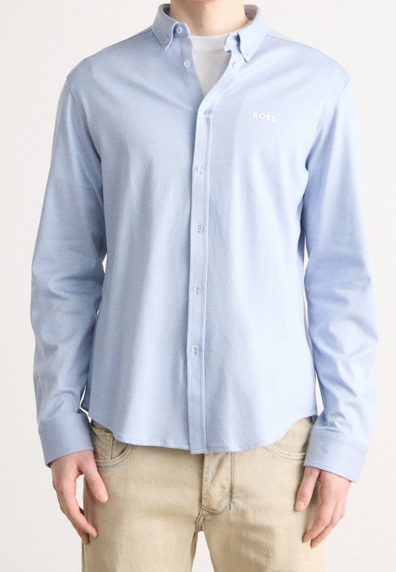 Ljusblå skjorta i bomull med knäppning, som har krage, långa ärmar och en logotyp på vänster bröst. Glatt material och figurnära design.