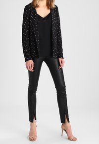 Svart kavaj med vitt diamantmönster, svart satin topp, smala svarta läderbyxor med slitsar vid fållen, och nakenfärgade sandaler med remmar.