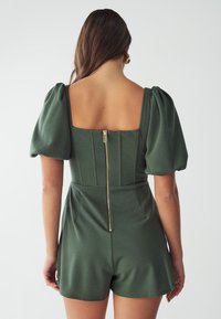 Romper ajusté vert avec de courtes manches bouffantes, dos carré et fermeture éclair. Il présente des panneaux structurés et un tissu lisse et texturé.