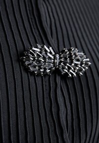 Svart plisserad tyg med en glänsande svart brosch med en rosettdesign, prydd med avlånga strass för extra textur och detalj.