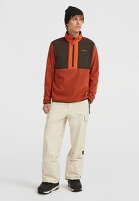Fleece-pullov i orange och brunt med halvdragkedja; kombinerat med krämfärgade cargobyxor och svarta stövlar. Texturerad tyg, praktisk design.