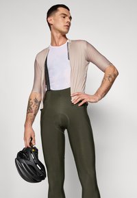 Équipement de cyclisme pour homme : maillot beige ajusté avec zip, sous-vêtement en maille blanche, cuissard forestier vert et casque de vélo noir.