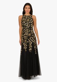 Žena v černých večerních šatech bez rukávů se zlatým květinovým vyšíváním a průhlednou vrstvenou sukní, stojící s uvolněnýma rukama.
