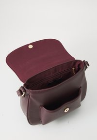 Sac à main en cuir bordeaux avec une fermeture à rabat, des accessoires dorés et une poche extérieure unique. L'intérieur comprend une poche zippée et un doublage doux.