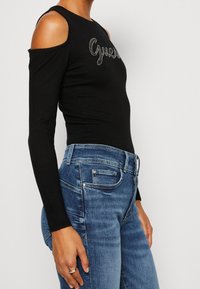 Haut noir à manches longues avec un design à épaules dénudées, arborant le texte argenté "Guess". Associé à un jean en denim bleu.