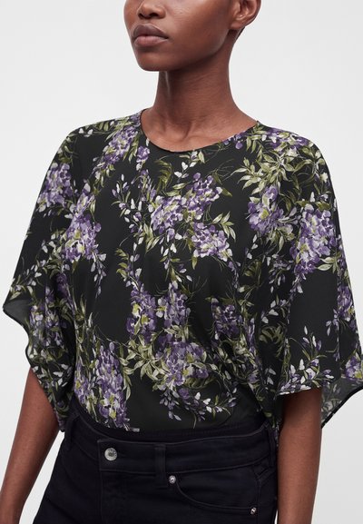 Femme portant un chemisier noir à imprimé floral violet et vert avec des manches courtes et fluides, rentrée dans un pantalon noir taille haute.