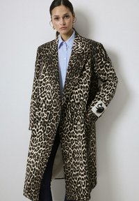 Femme portant un long manteau en imprimé léopard sur une chemise boutonnée bleu clair, posant avec une main dans la poche du manteau devant un fond uni.