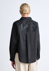 Black shirt featuring vertical stripes, a collar, and a relaxed fit. Textured upper panel with subtle sheen, long sleeves, and curved hem.