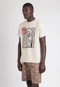 Camiseta de algodón color crema con un estampado gráfico en negro y rojo de un personaje sosteniendo un pez, combinada con pantalones cortos marrones con un patrón.