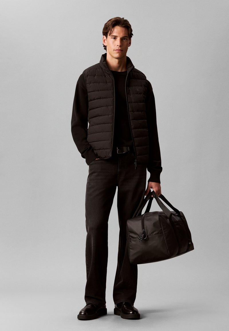 Homme portant une doudoune noire sans manches, une chemise noire à manches longues, un pantalon noir et des chaussures noires, tenant un sac de sport noir dans sa main droite.
